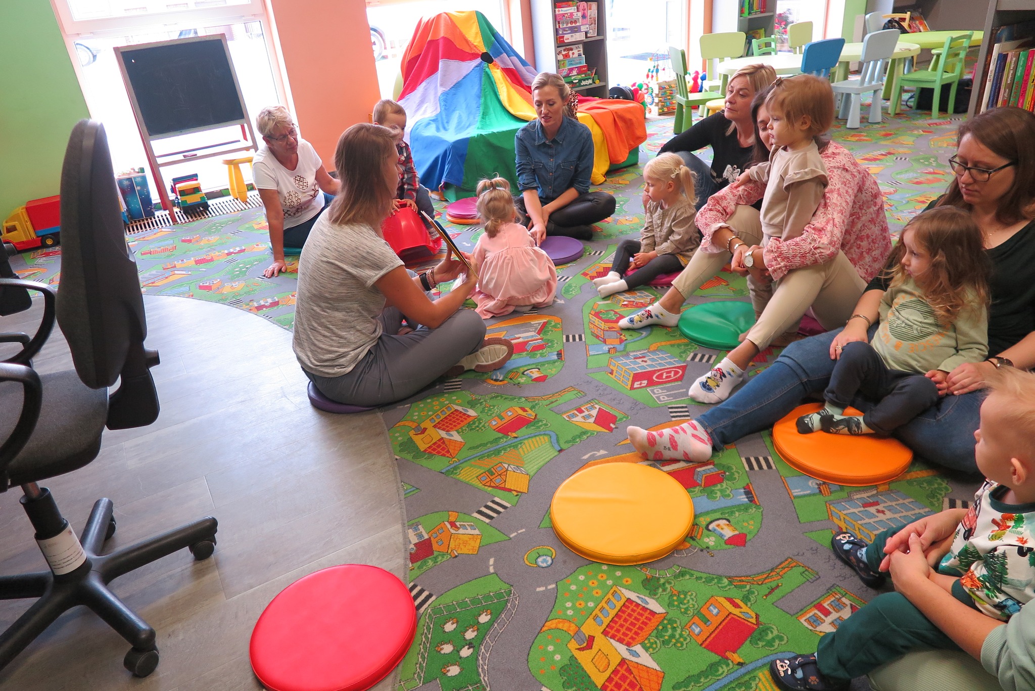 Dzieci z rodzicem siedzą na dywanie w oddziele dla dzieci z bibliotekarką