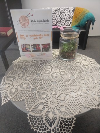 haftowany obrus na stoliku na którym stoi plakat klubu rękodzieła i kwiat w szklanym słoju