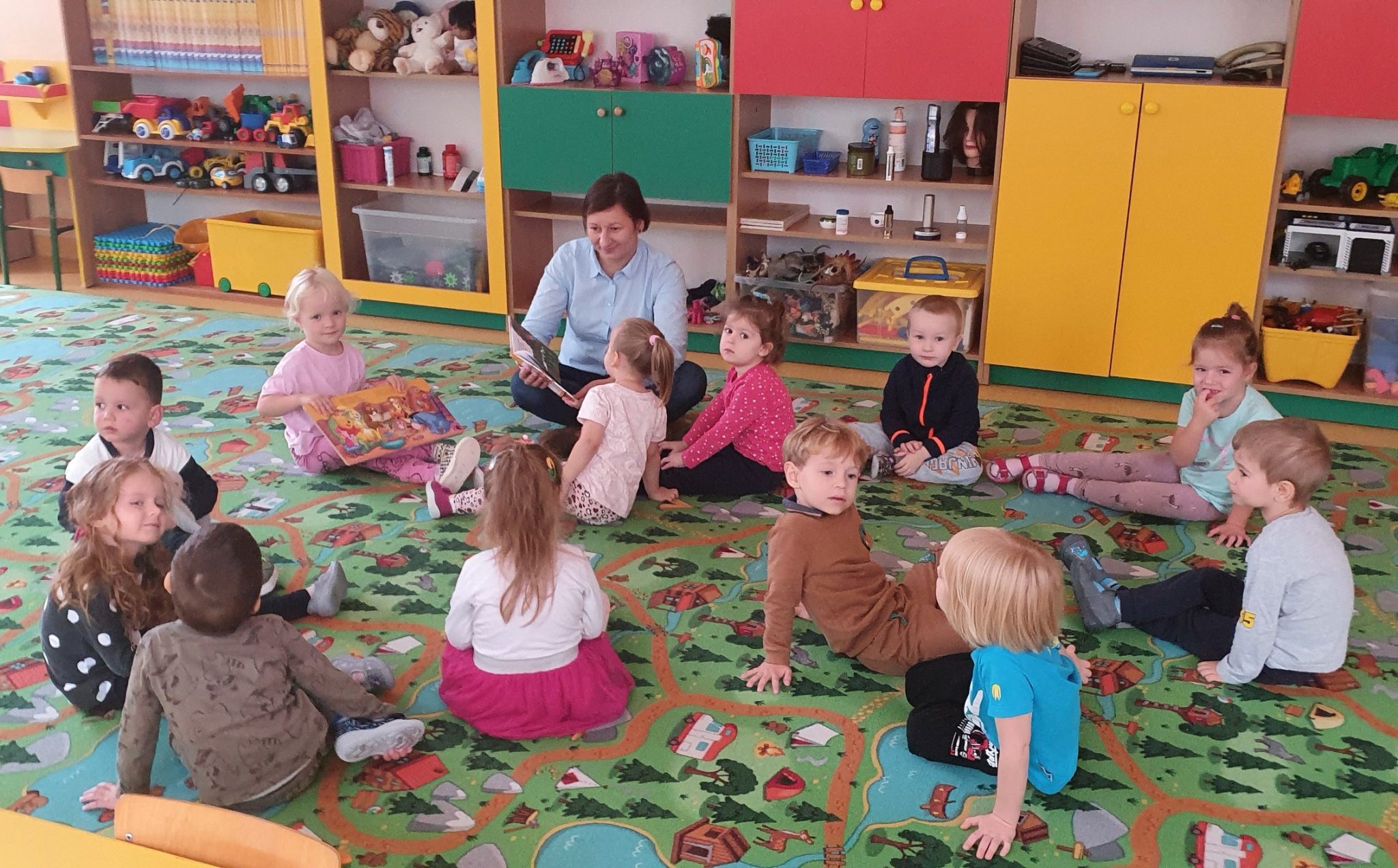 dzieci siedzą na dywanie w sali przedszkolnej i słuchają czytanej przez bibliotekarkę książki