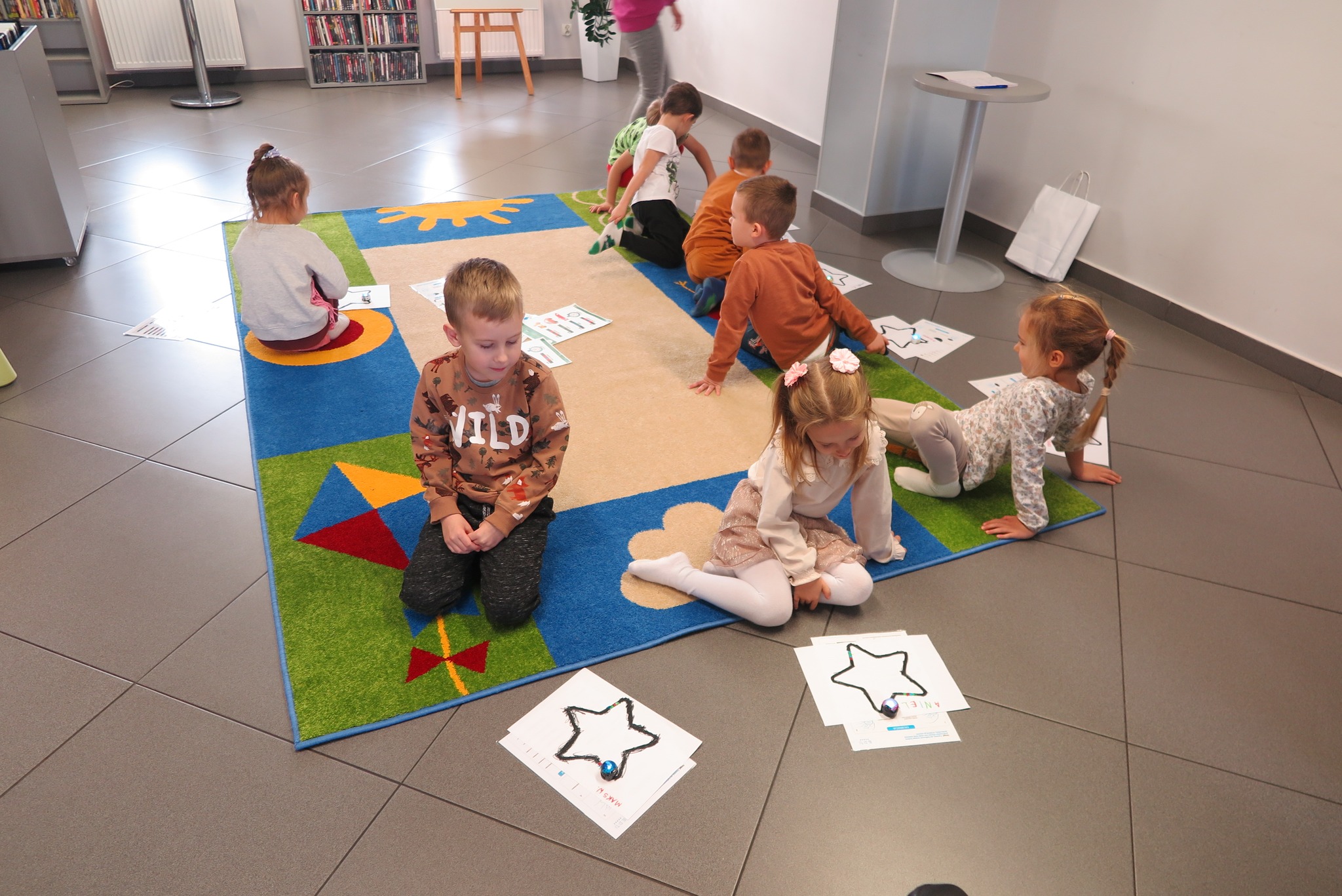 dzieci puszczają ozoboty po trasie w kształcie gwiazdy siedząc na dywanie w mediatece