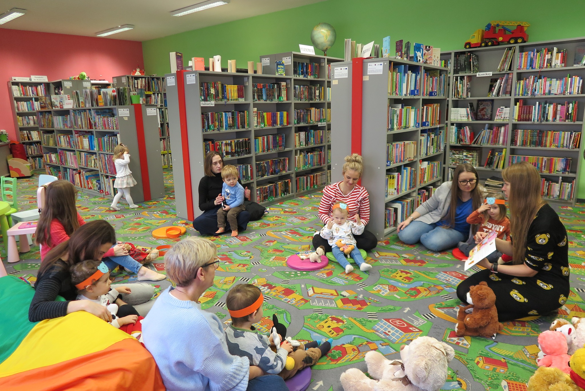 dzieci z klubu malucha siedzą z rodzicem na dywanie w oddziale dla dzieci  gdzie bibliotekarka czyta książkę w otoczeniu pluszowych misiów