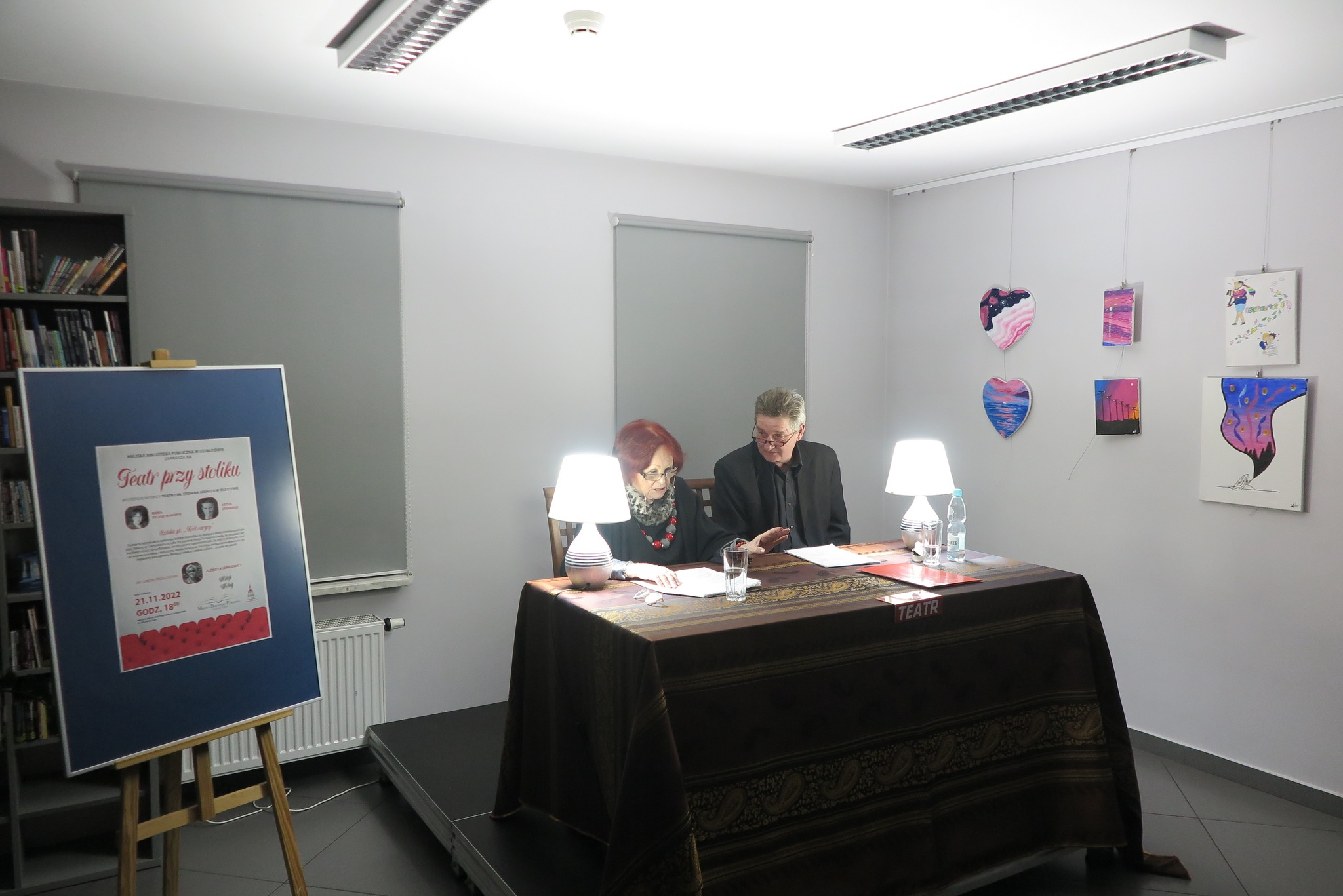 Irena Telesz-Burczyk i Artur Steranko siedzą za stolikiem przykrytym brązowym obrusem  na którym stoją dwie lampki nocne
