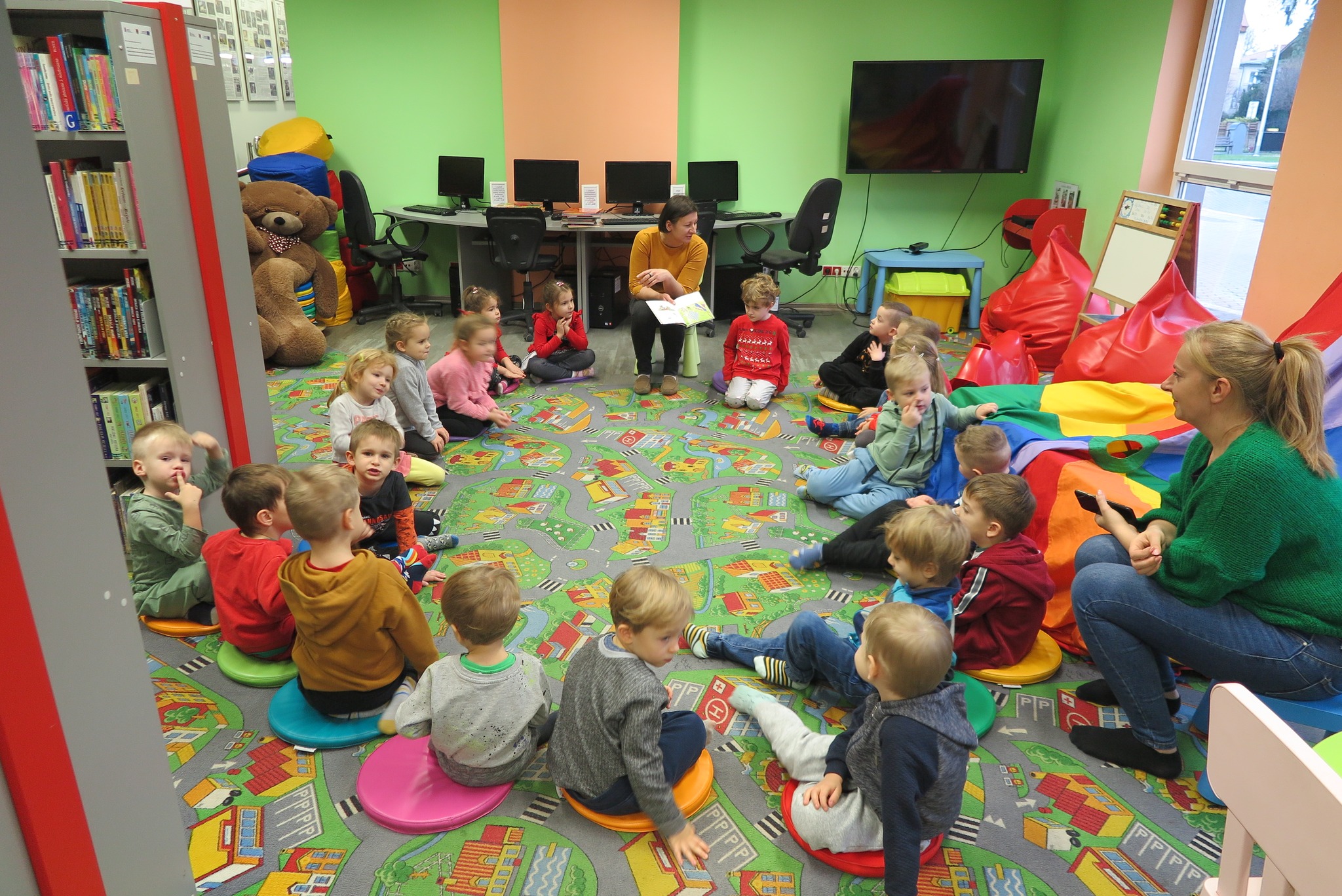 dzieci z przedszkola nr 5 siedzą na dywanie w oddziale dla dzieci i słuchają bajki czytanej przez bibliotekarkę 