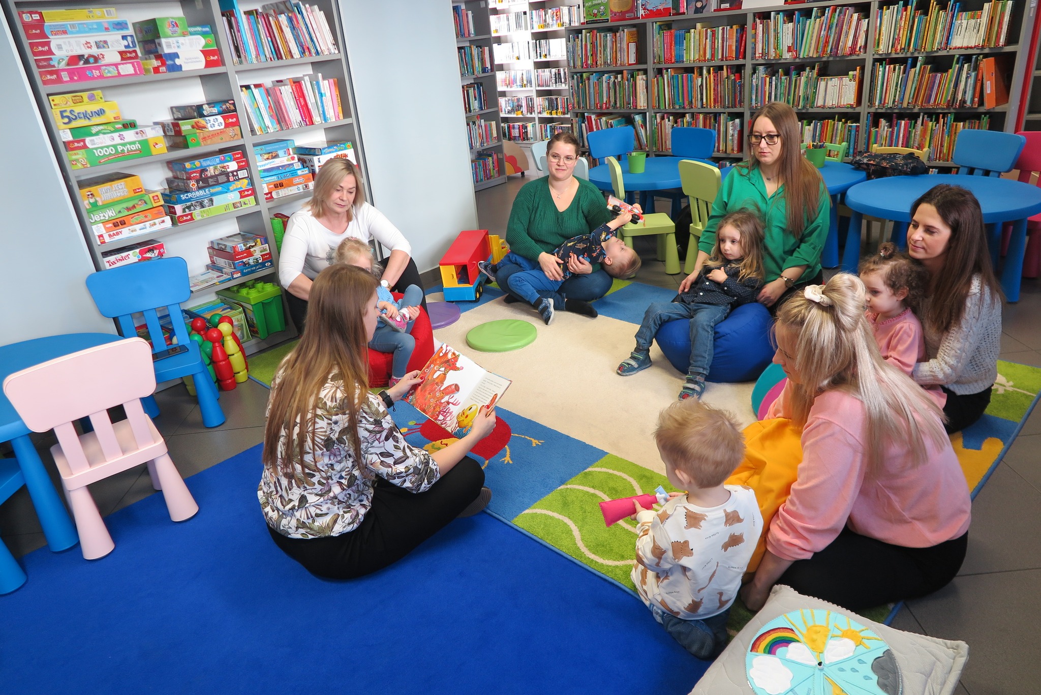 Dzieci z rodzicami siedzą na dywanie w Oddziale dla dzieci i słuchają czytanej przez bibliotekarkę książki