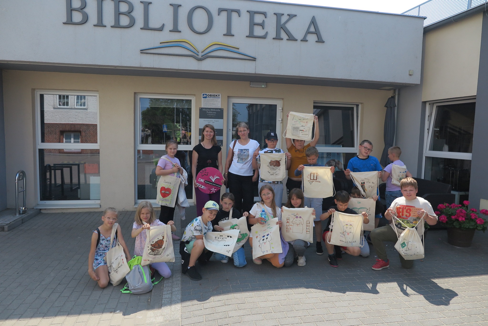 Członkowie klubu DKK SP1 pokazują pomalowane torby, stoją przed biblioteką