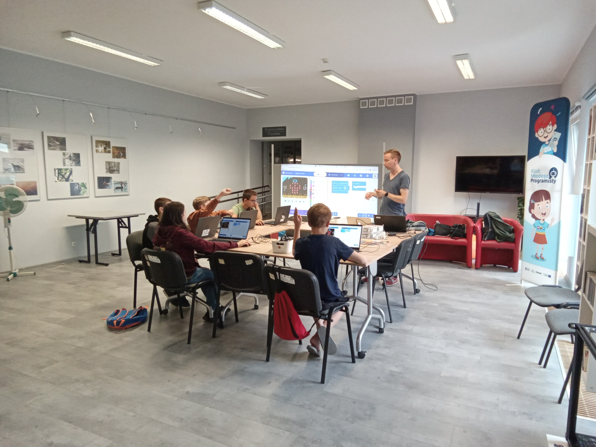 uczestnicy warsztatów Klubu Młodego Programisty siedzą z laptopami przy stole, w tle na ekranie wyświetlany jest obraz z projektora