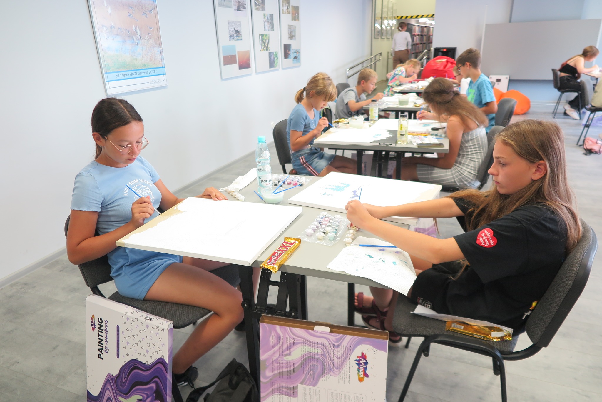 uczestnicy warsztatów malowania po numerach siedzą przy dwuosobowych stolikach, na których leżą płótna i farby, malują 