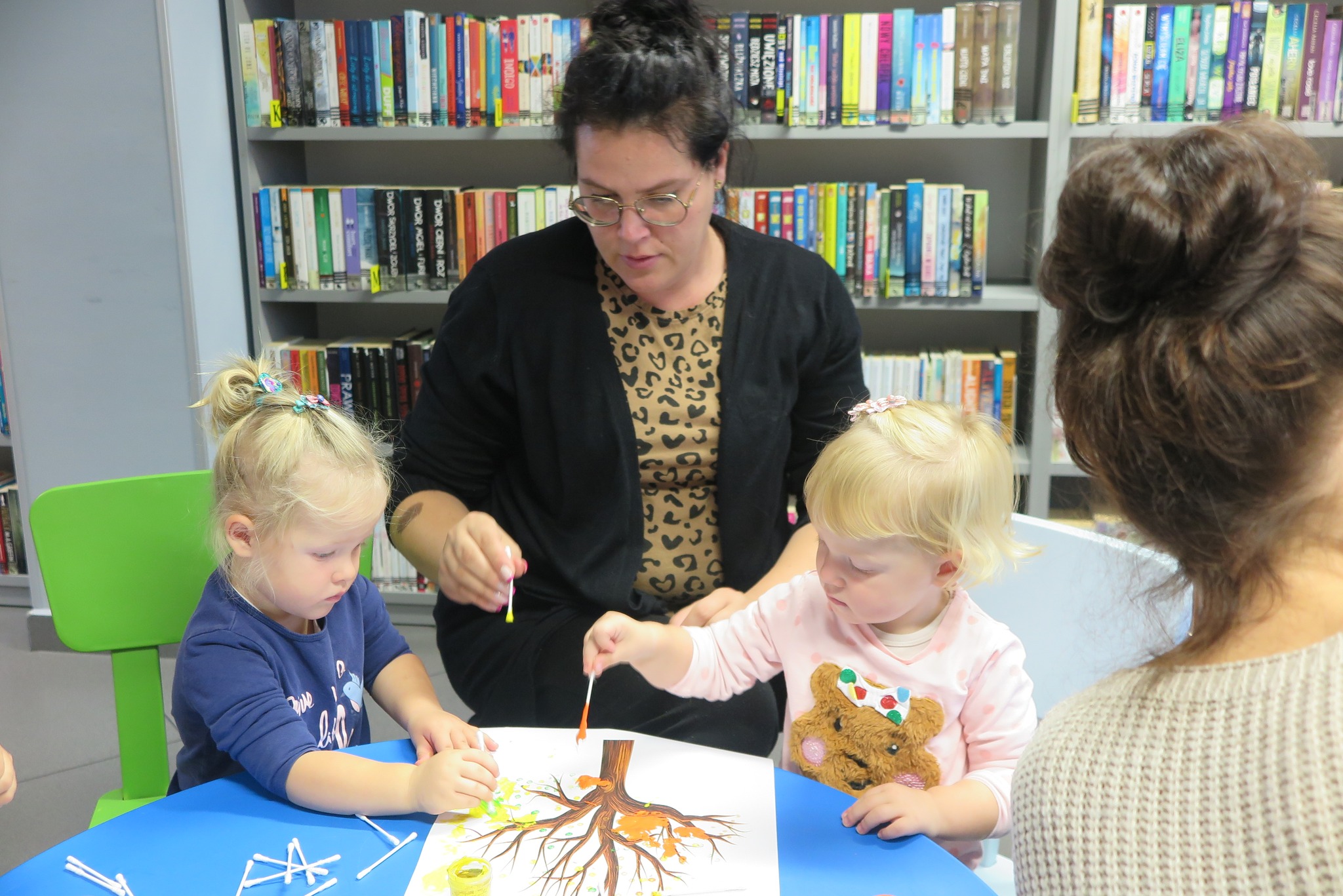 dzieci z rodzicami podczas spotkania klubu malucha malują liście na kartce z rysunkiem drzewa