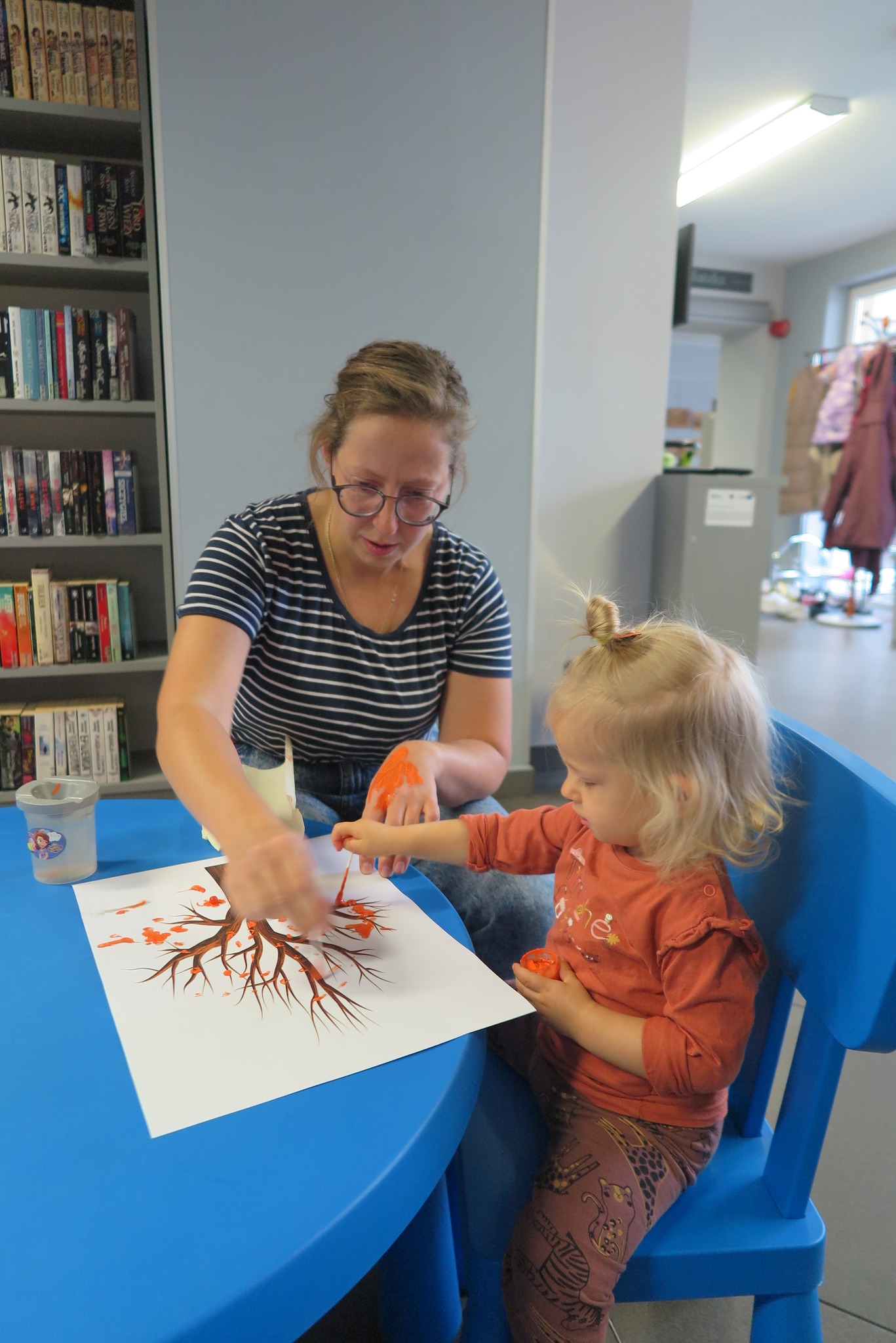 dzieci z rodzicami podczas spotkania klubu malucha malują liście na kartce z rysunkiem drzewa