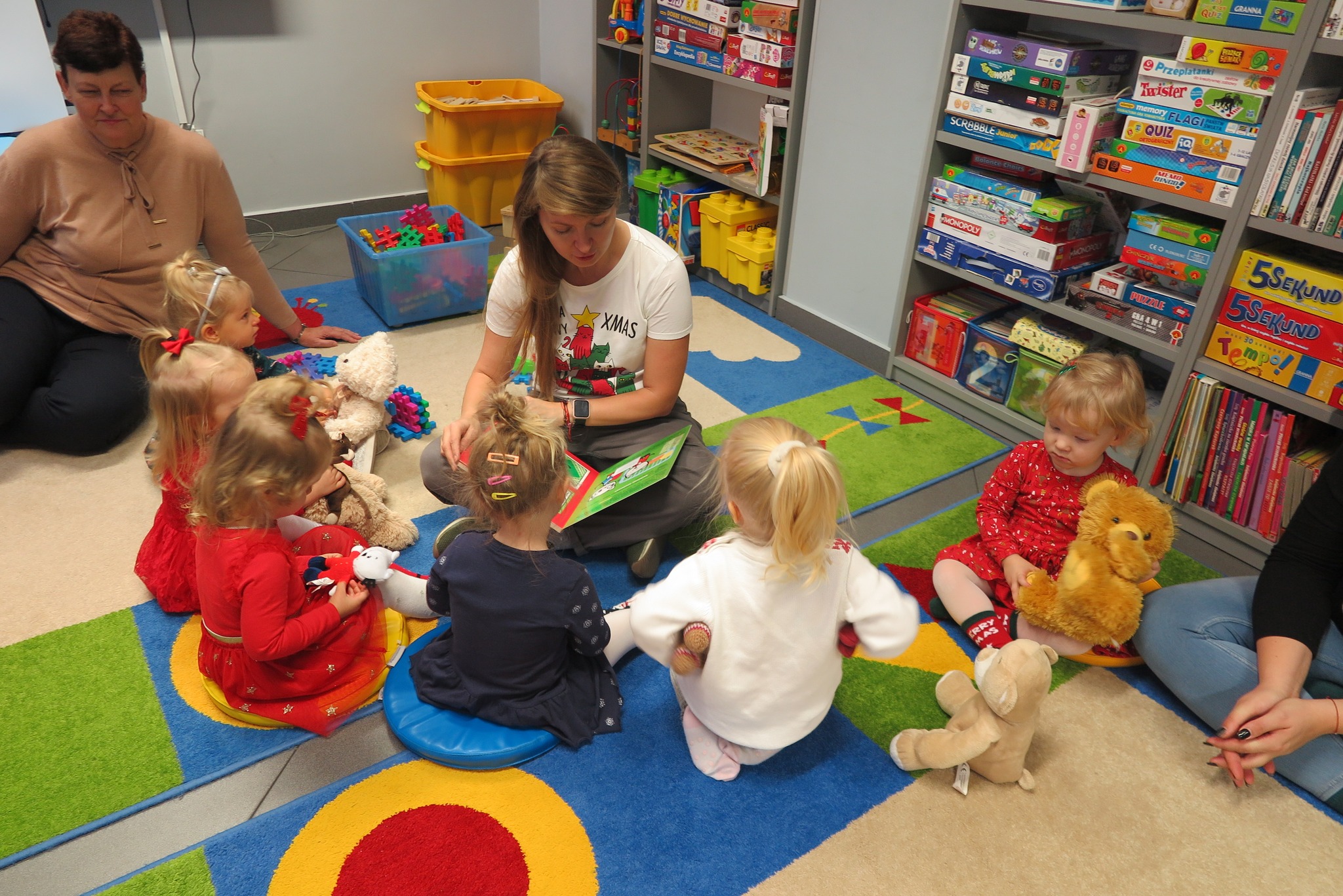 dzieci z klubu malucha słuchają czytanej książki siedząc na dywanie w oddziale dla dzieci
