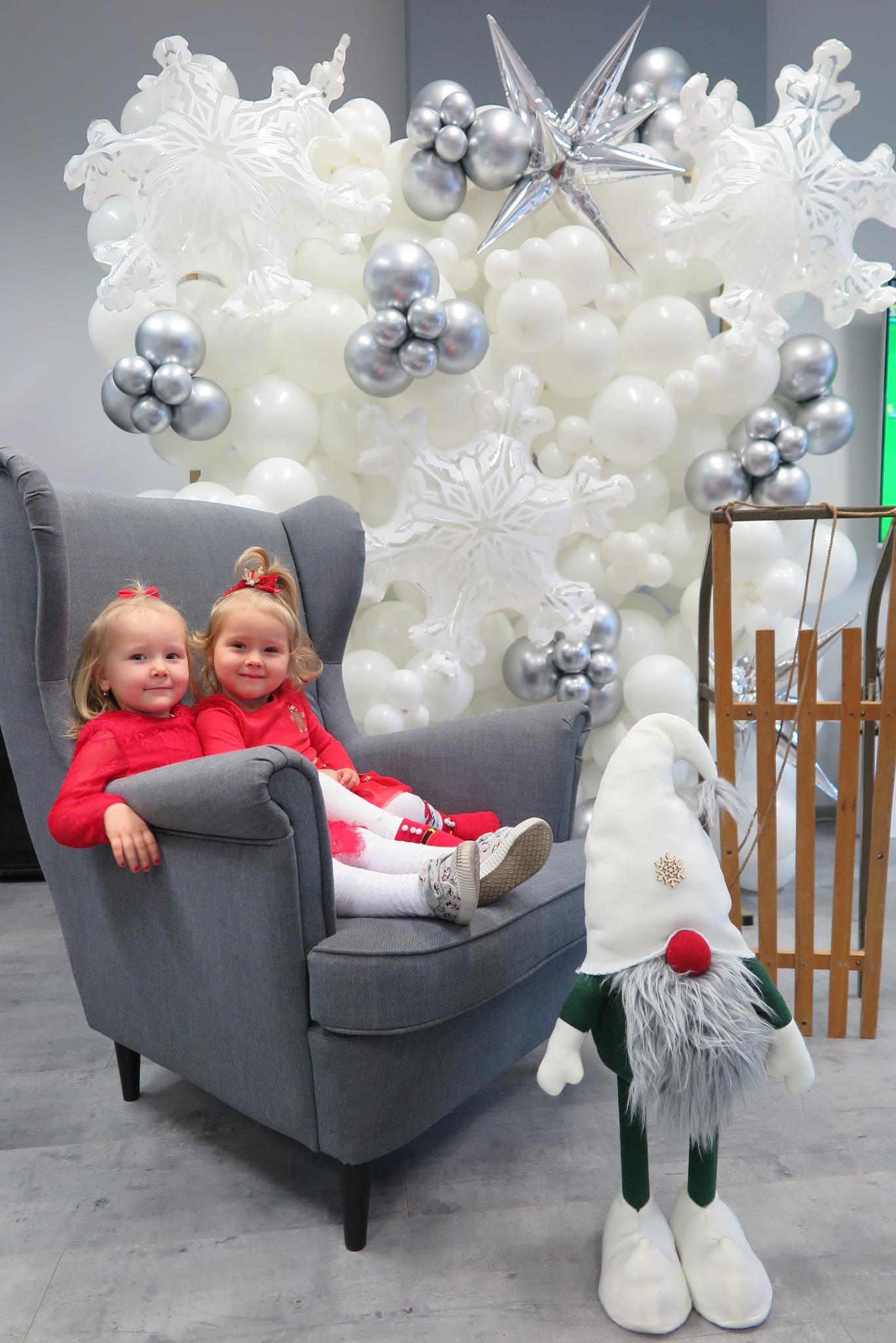 dwójka dzieci siedzi w szarym fotelu, za nimi stoi ścianka wykonana z balonów, po prawej stoją sanki i biało-zielony skrzat 